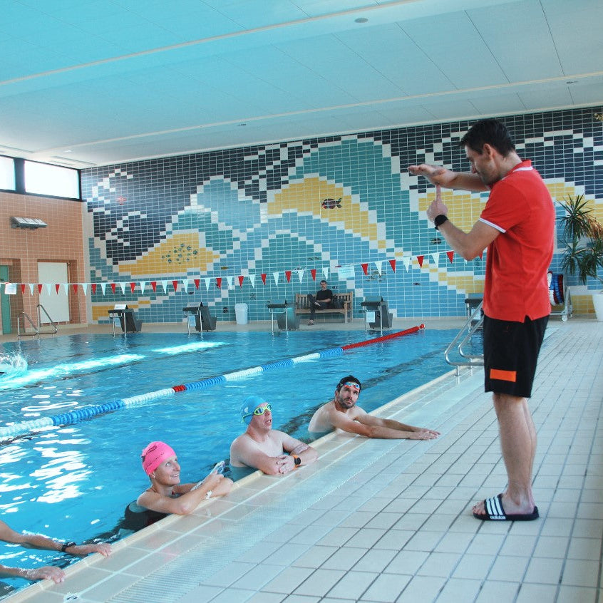 Ein Schwimmtrainer steht am Beckenrand und erklärt 4 Schwimmern die Schwimmtechnik