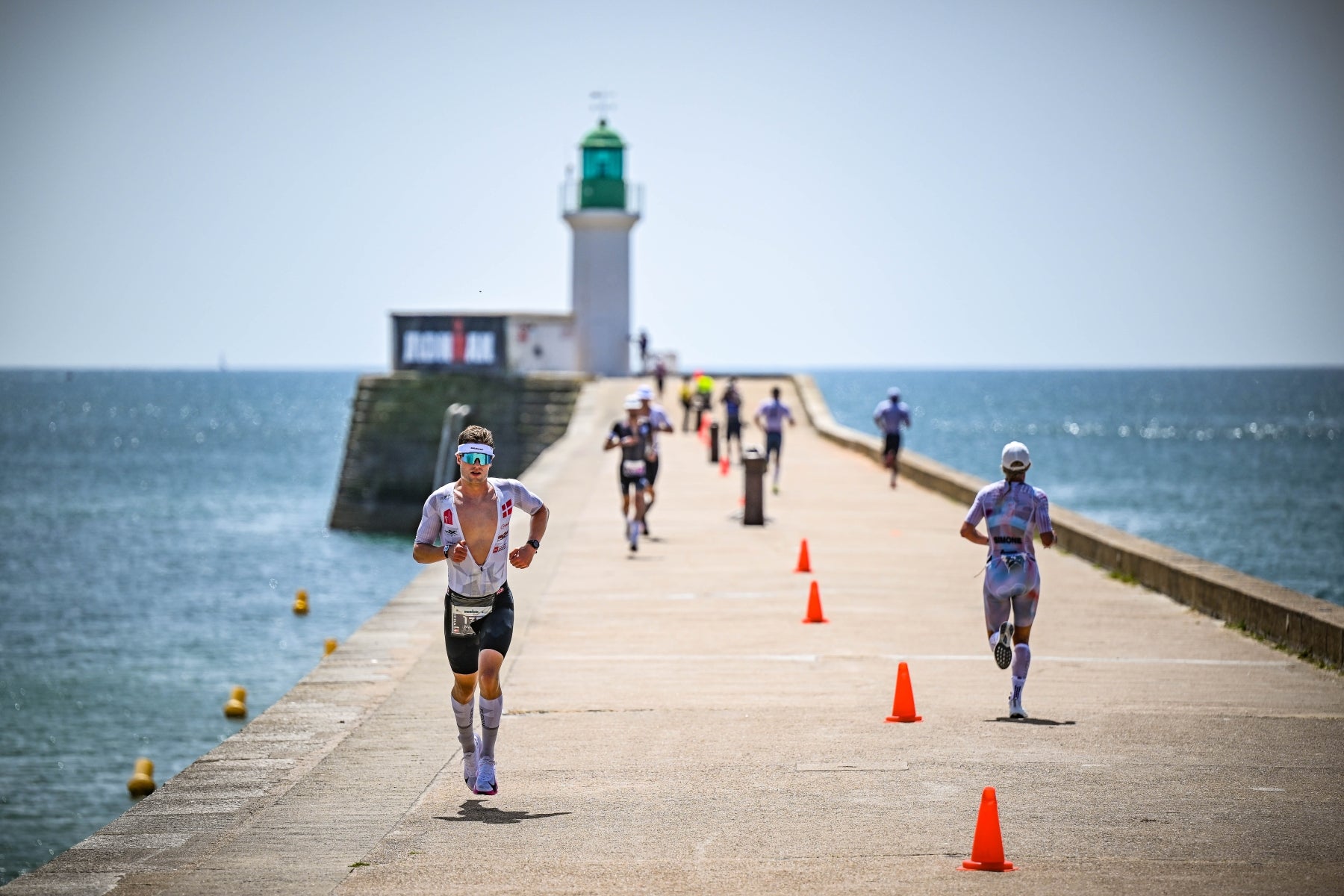 Triathleten auf der Laufstrecke des Ironman Les Sables d'Olonne-Vendee laufen auf der Hafenmole auf einen Leuchtturm zu, wo sich der Wendepunkt befindet